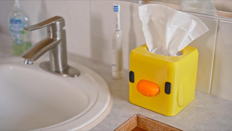 Pu&ograve; includere: Scatola di fazzoletti di plastica gialla a forma di pulcino dei cartoni animati con un becco arancione e occhi neri. La scatola &egrave; piena di fazzoletti bianchi.