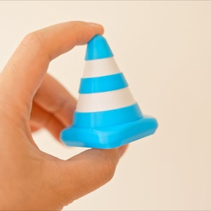 Op de afbeelding: Een blauw en wit verkeerskegel speelgoed in een hand gehouden. De kegel heeft drie witte strepen en een blauwe basis.