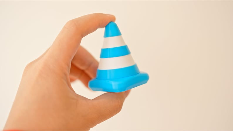 Op de afbeelding: Een blauw en wit verkeerskegel speelgoed in een hand gehouden. De kegel heeft drie witte strepen en een blauwe basis.