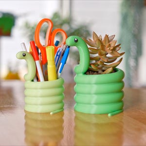 May include: Two whimsical, snake-shaped desk organizers. One holds pens, markers, and scissors. The other is a small planter with a succulent. Both are light green and sit on a wooden surface.