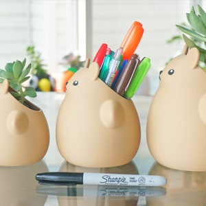 May include: Three ceramic animal-shaped planters, two with small succulents and one with a collection of pens. The planters are beige and have a cartoonish design. A black Sharpie marker is in the foreground.