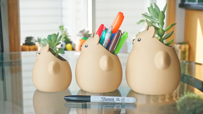 May include: Three ceramic animal-shaped planters, two with small succulents and one with a collection of pens. The planters are beige and have a cartoonish design. A black Sharpie marker is in the foreground.
