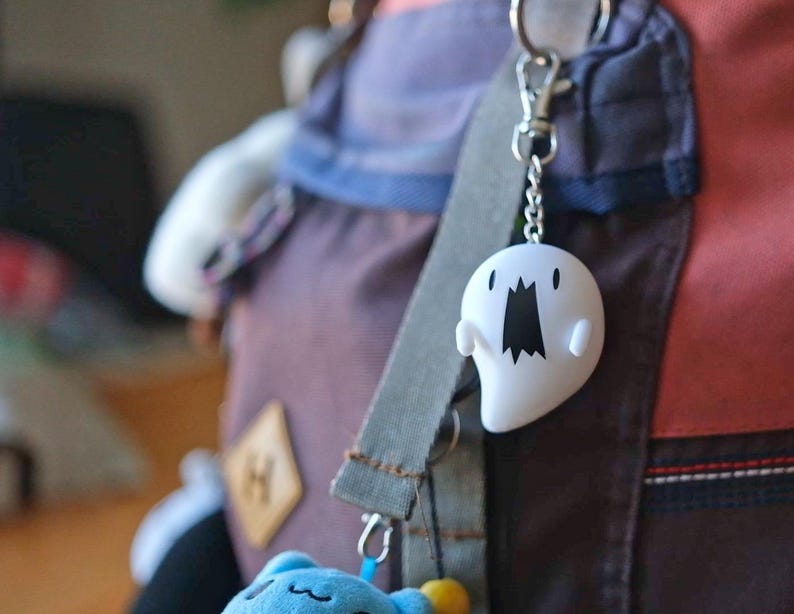 May include: A white ghost-shaped keychain with a black mouth and eyes hangs from a silver chain. The keychain is attached to a backpack with a gray strap. A blue plush toy is visible at the bottom.