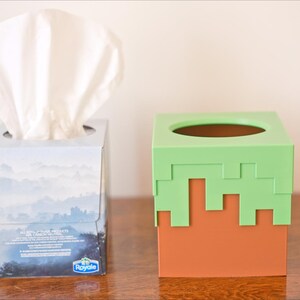 A cube tissue box cover inspired by the dirt grass block from the popular video game, Minecraft, is on a wooden counter next to the tissue box that it can cover. It has bright green pixelated grass at the top half and the bottom half is brown.