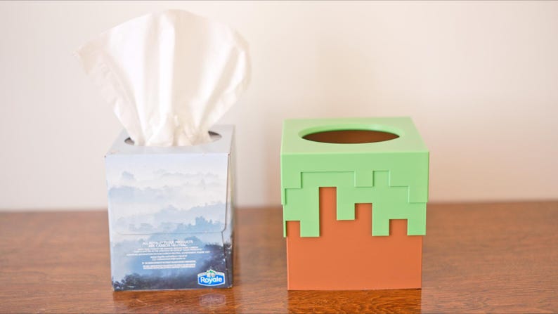 A cube tissue box cover inspired by the dirt grass block from the popular video game, Minecraft, is on a wooden counter next to the tissue box that it can cover. It has bright green pixelated grass at the top half and the bottom half is brown.
