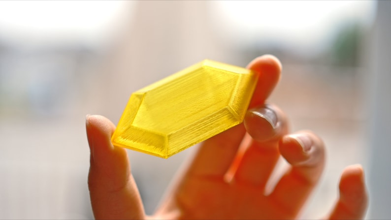May include: A yellow, faceted, plastic gem. The gem is held in a hand.