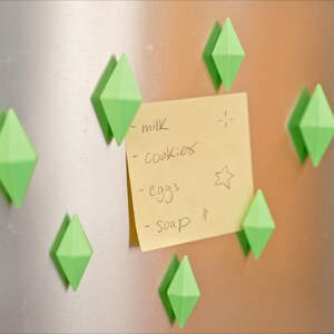 May include: A yellow sticky note with a handwritten grocery list on a stainless steel refrigerator. The list includes milk, cookies, eggs, and soap. The note is held in place by several green diamond-shaped magnets.