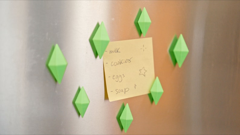 May include: A yellow sticky note with a handwritten grocery list on a stainless steel refrigerator. The list includes milk, cookies, eggs, and soap. The note is held in place by several green diamond-shaped magnets.