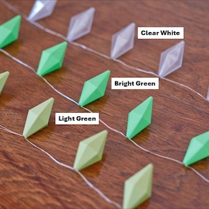 May include: A string of decorative lights with diamond-shaped ornaments in clear white, bright green, and light green. The lights are arranged on a wooden surface, with the text "Clear White", "Bright Green", and "Light Green" visible.