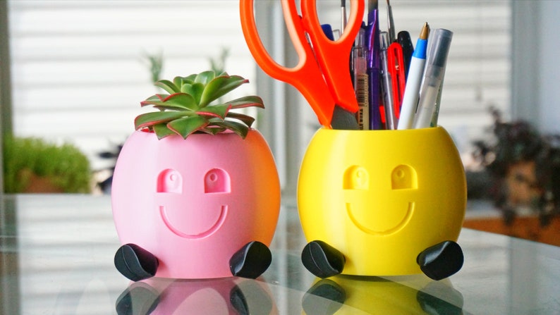 May include: Two 3D printed smiley face planters, one pink with a succulent plant and the other yellow with a set of pens and scissors.