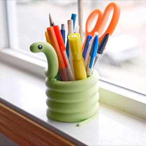 May include: A light green, snake-shaped desk organizer filled with pens, pencils, and orange scissors. The organizer sits on a white windowsill with a view of the outside.