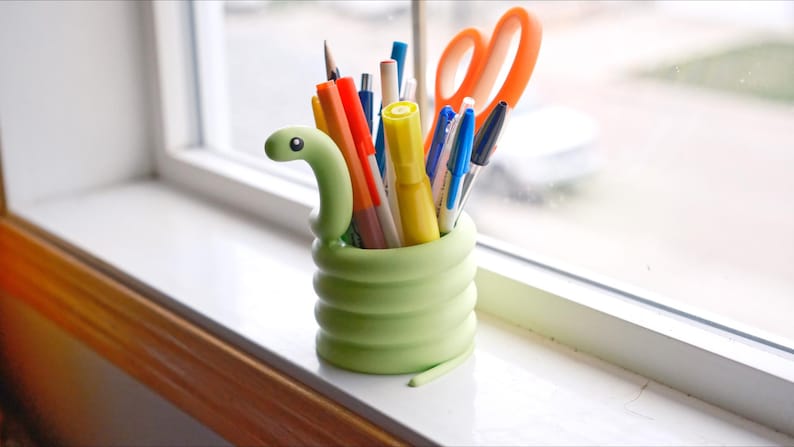 May include: A light green, snake-shaped desk organizer filled with pens, pencils, and orange scissors. The organizer sits on a white windowsill with a view of the outside.