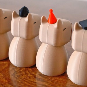 May include: Four brown ceramic figurines with different coloured hats. The hats are yellow, black, red, and black. The figurines have a simple design and are standing on a wooden surface.