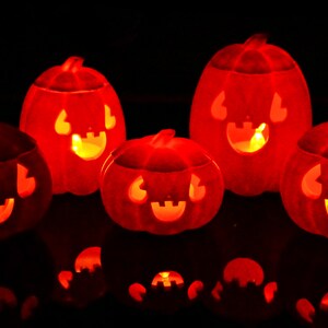 May include: Five orange jack-o'-lantern pumpkins with carved faces are lit from within, casting a warm glow. The pumpkins are arranged on a dark surface, creating a festive Halloween scene.