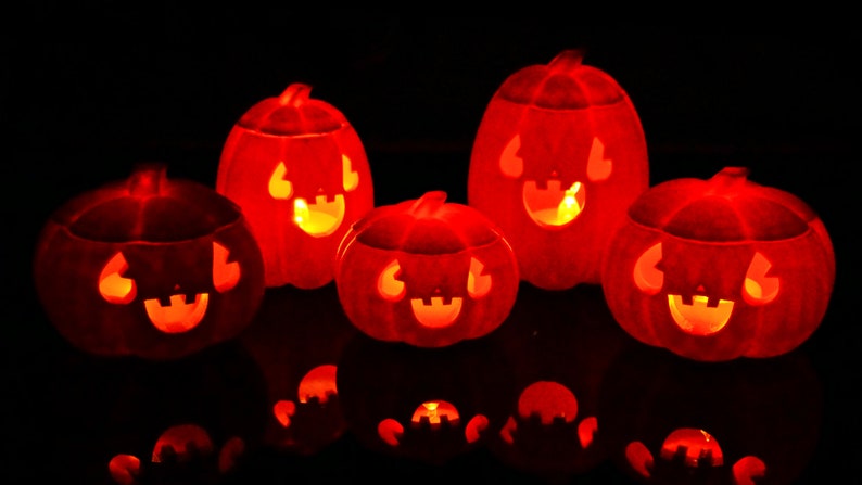 May include: Five orange jack-o'-lantern pumpkins with carved faces are lit from within, casting a warm glow. The pumpkins are arranged on a dark surface, creating a festive Halloween scene.