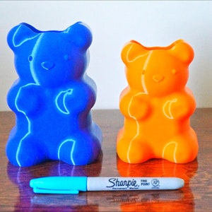 Puede incluir: Dos porta l&aacute;pices con forma de osito de goma, uno azul y otro naranja, sobre una superficie de madera. Un marcador azul est&aacute; delante de los porta l&aacute;pices.