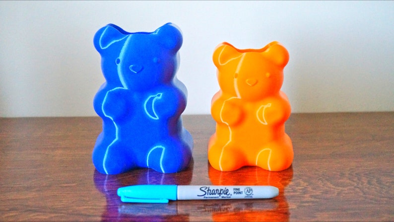 Puede incluir: Dos porta l&aacute;pices con forma de osito de goma, uno azul y otro naranja, sobre una superficie de madera. Un marcador azul est&aacute; delante de los porta l&aacute;pices.