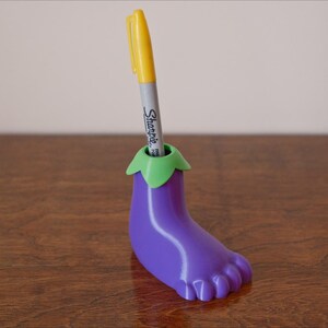 May include: Purple eggplant-shaped pen holder with a green top and a yellow pen sticking out of the top. The pen holder is sitting on a wooden surface.