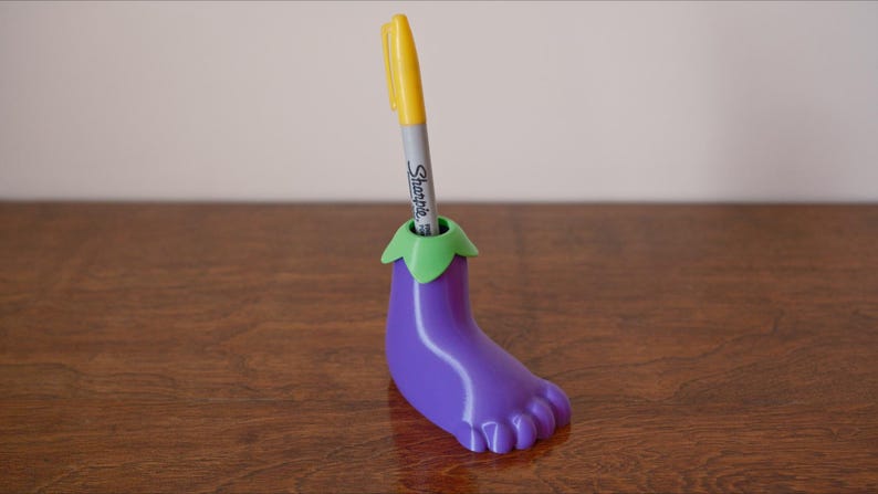 May include: Purple eggplant-shaped pen holder with a green top and a yellow pen sticking out of the top. The pen holder is sitting on a wooden surface.