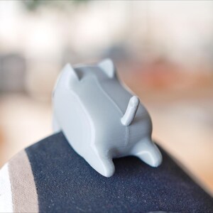 May include: A small, grey, cat-shaped ornament with a tail and four legs. The ornament is resting on a dark surface. The cat figurine is a decorative item.