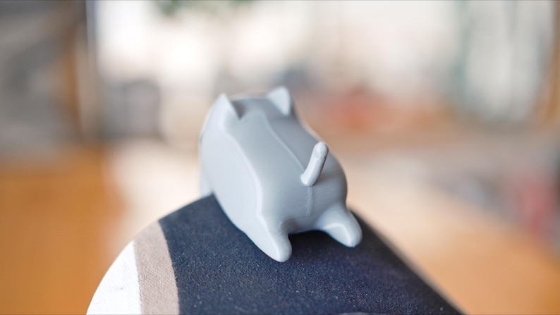 May include: A small, grey, cat-shaped ornament with a tail and four legs. The ornament is resting on a dark surface. The cat figurine is a decorative item.
