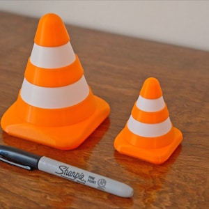 Op de afbeelding: Twee oranje en witte verkeerskegels, een groter dan de andere, op een houten oppervlak. Een zwarte stift ligt op het oppervlak.