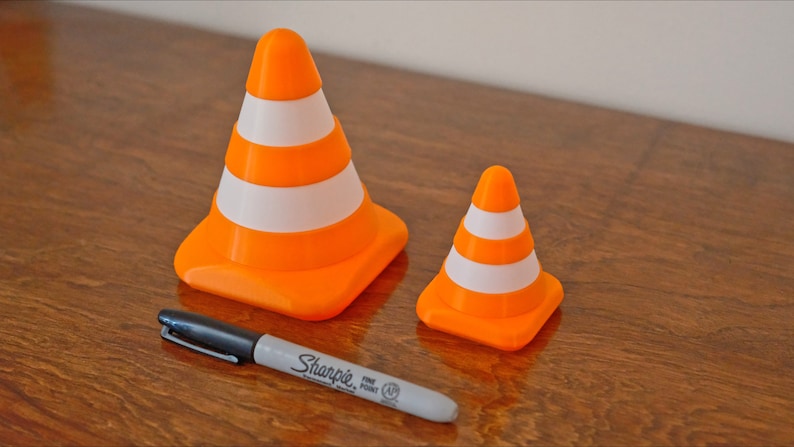 Op de afbeelding: Twee oranje en witte verkeerskegels, een groter dan de andere, op een houten oppervlak. Een zwarte stift ligt op het oppervlak.