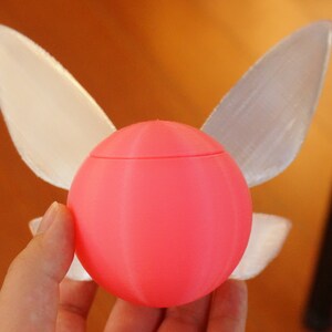 Puede incluir: Una esfera rosa impresa en 3D con dos alas blancas transl&uacute;cidas unidas a los lados. La esfera tiene una superficie lisa y redondeada.