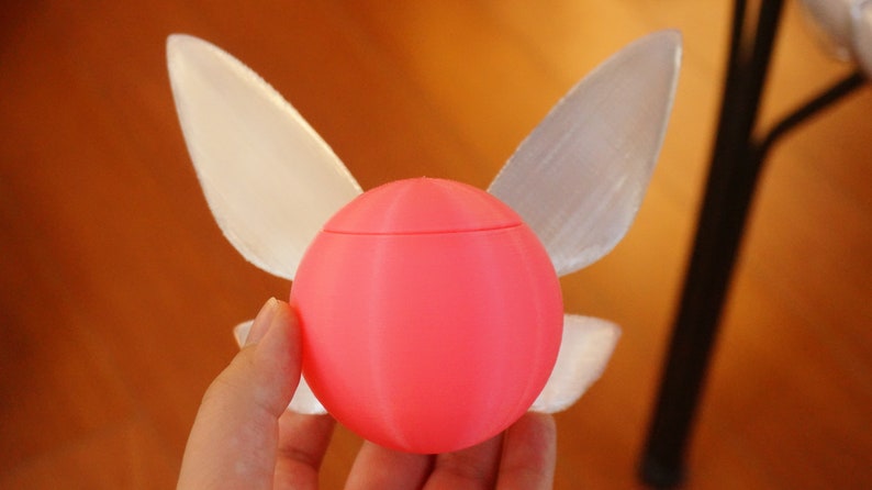 Puede incluir: Una esfera rosa impresa en 3D con dos alas blancas transl&uacute;cidas unidas a los lados. La esfera tiene una superficie lisa y redondeada.