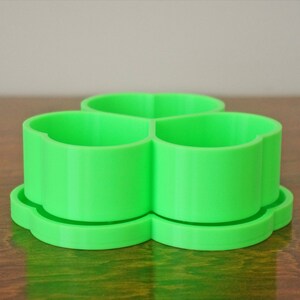 May include: A bright green, three-sectioned container, likely 3D-printed, sits on a matching base. The container's design features rounded edges and a clover-like shape. The object is on a wooden surface.
