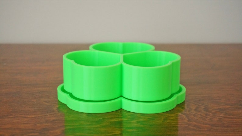 May include: A bright green, three-sectioned container, likely 3D-printed, sits on a matching base. The container's design features rounded edges and a clover-like shape. The object is on a wooden surface.