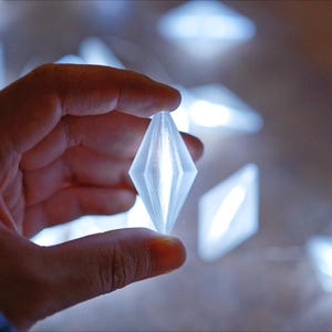 May include: A hand holding a glowing, diamond-shaped light. Several similar lights are out of focus in the background. The lights appear to be part of a decorative string, emitting a soft, cool white glow.