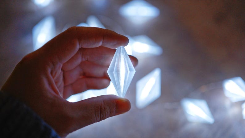 May include: A hand holding a glowing, diamond-shaped light. Several similar lights are out of focus in the background. The lights appear to be part of a decorative string, emitting a soft, cool white glow.