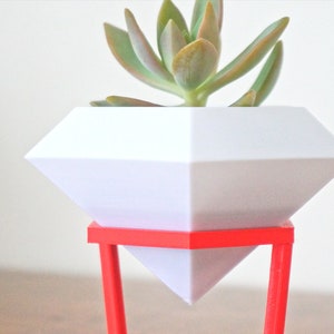 May include: A white diamond-shaped planter with a succulent plant inside, sitting on a red metal stand.
