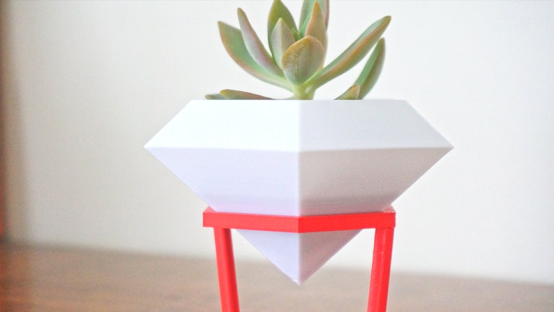 May include: A white diamond-shaped planter with a succulent plant inside, sitting on a red metal stand.