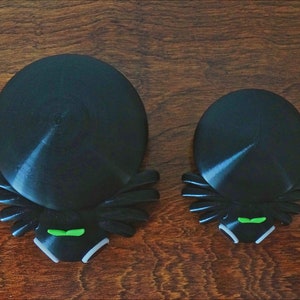 May include: Two black spider-shaped bottle stoppers with green eyes. The larger stopper is about 4 inches in diameter and the smaller stopper is about 3 inches in diameter.