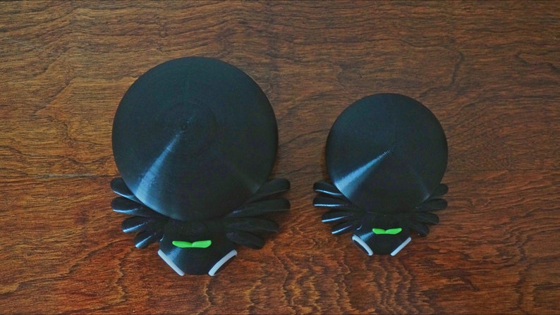 May include: Two black spider-shaped bottle stoppers with green eyes. The larger stopper is about 4 inches in diameter and the smaller stopper is about 3 inches in diameter.