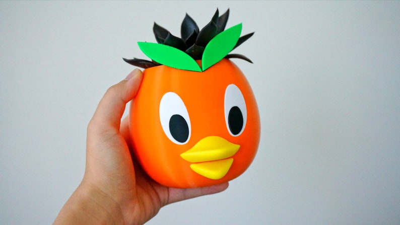 May include: Orange ceramic planter shaped like a duck with black eyes, a yellow beak, and green leaves on top.