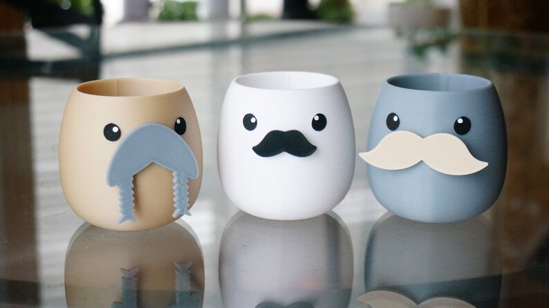 May include: Three ceramic cups with different facial expressions. The first cup is brown with a gray walrus-like mustache. The second cup is white with a black mustache. The third cup is gray with a cream-colored mustache.