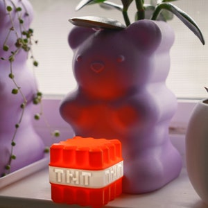 Op de afbeelding: Een paarse keramische berenpot met een groene vetplant erin. De pot staat op een vensterbank naast een rood en wit blokje met het woord "TNT" erop.