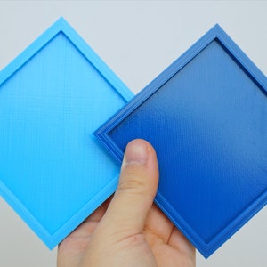 K&ouml;nnte beinhalten: Zwei quadratische Tabletts, eines hellblau und eines dunkelblau, mit einem erh&ouml;hten Rand um die Kanten. Die Tabletts werden in einer Hand gehalten.
