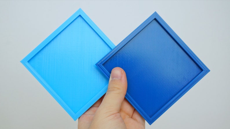 K&ouml;nnte beinhalten: Zwei quadratische Tabletts, eines hellblau und eines dunkelblau, mit einem erh&ouml;hten Rand um die Kanten. Die Tabletts werden in einer Hand gehalten.