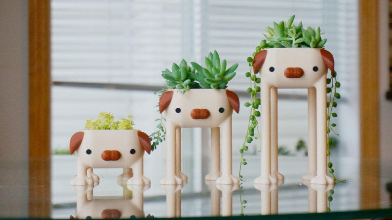 May include: Three white ceramic planters shaped like dogs with brown ears and noses. Each planter has a different size and holds a variety of green succulents.