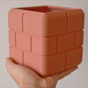 Pu&ograve; includere: Un vaso da fiori in ceramica color terracotta a forma di pila di mattoni. Il vaso ha una forma quadrata e una superficie testurizzata.