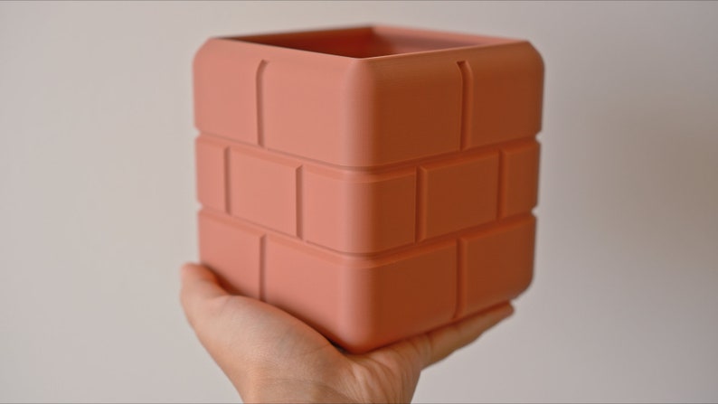Pu&ograve; includere: Un vaso da fiori in ceramica color terracotta a forma di pila di mattoni. Il vaso ha una forma quadrata e una superficie testurizzata.