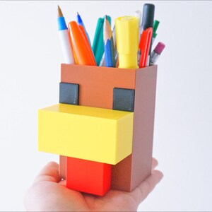 K&ouml;nnte beinhalten: Ein brauner, gelber und roter Minecraft-Stiftbeh&auml;lter in Form eines Huhns. Der Beh&auml;lter hat einen gelben Schnabel, einen roten Kamm und schwarze Augen.