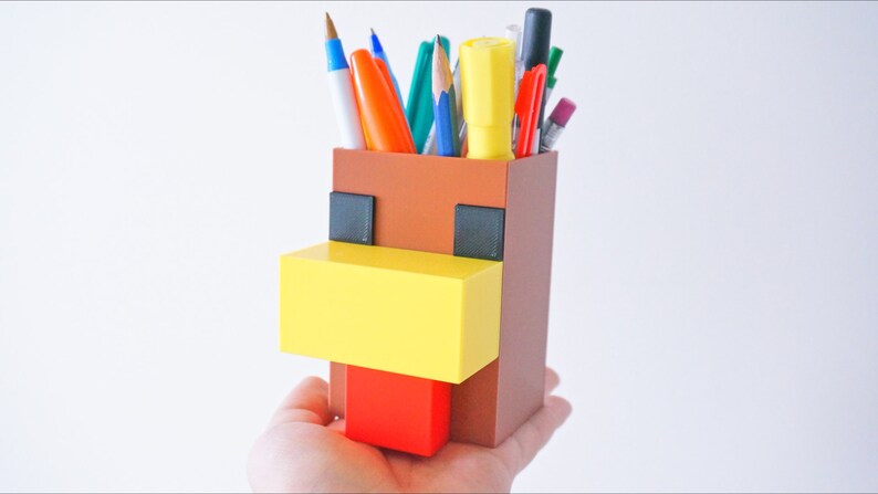 K&ouml;nnte beinhalten: Ein brauner, gelber und roter Minecraft-Stiftbeh&auml;lter in Form eines Huhns. Der Beh&auml;lter hat einen gelben Schnabel, einen roten Kamm und schwarze Augen.