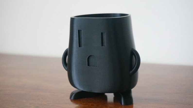 Op de afbeelding: Een zwarte 3D-geprinte potloodhouder in de vorm van een cartoonfiguur met een verraste uitdrukking. Het personage heeft een stopcontact voor een gezicht en kleine benen.