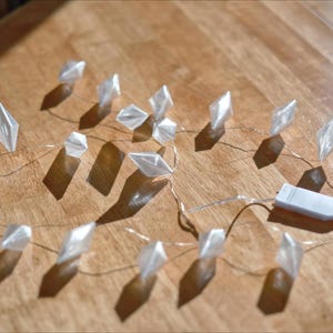 May include: A string of clear, diamond-shaped LED lights on a thin wire, with a white battery pack. The lights are arranged on a wooden surface, casting shadows. The lights are a decorative element.