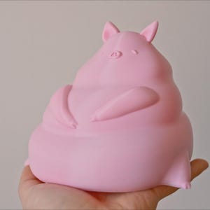 May include: A pink, 3D printed pig figurine with a rounded, plump body and small ears. The pig is sitting with its arms crossed in front of it.